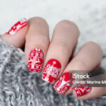 A festive red Christmas manicure featuring intricate winter designs. One nail showcases a white reindeer silhouette, while others display delicate snowflakes and a Norwegian sweater pattern in white. The vibrant red base color enhances the holiday spirit, and the detailed white patterns evoke cozy winter vibes. The combination of reindeer, snowflakes, and the classic Norwegian design creates a cheerful and stylish holiday look.