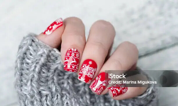 A festive red Christmas manicure featuring intricate winter designs. One nail showcases a white reindeer silhouette, while others display delicate snowflakes and a Norwegian sweater pattern in white. The vibrant red base color enhances the holiday spirit, and the detailed white patterns evoke cozy winter vibes. The combination of reindeer, snowflakes, and the classic Norwegian design creates a cheerful and stylish holiday look.