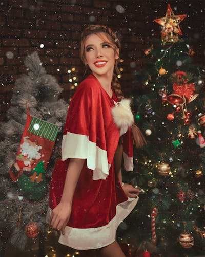 "Elegant woman in a short santa dress embodying a celebrity Christmas look. She wears glamorous makeup with a bold red lip and classic winged eyeliner, her hair styled in soft waves adorned with subtle sparkly accessories. The festive background features warm Christmas lights, a decorated tree, and an upscale holiday ambiance, capturing a chic and sophisticated holiday style."