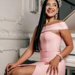 miss universe is sitting on the table. she is wearing pink color gown and a crown . she is looking beautiful and attractive. Her smile is catching the limelight.