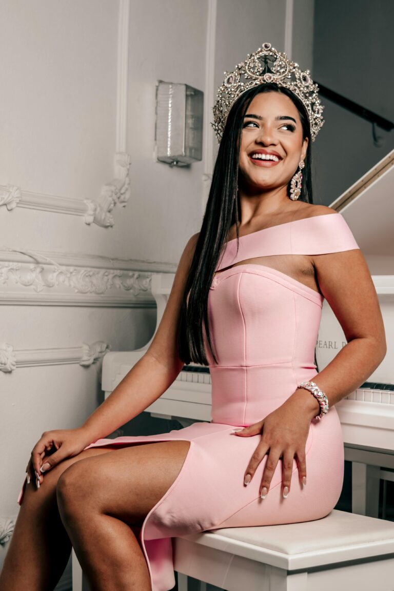 miss universe is sitting on the table. she is wearing pink color gown and a crown . she is looking beautiful and attractive. Her smile is catching the limelight.